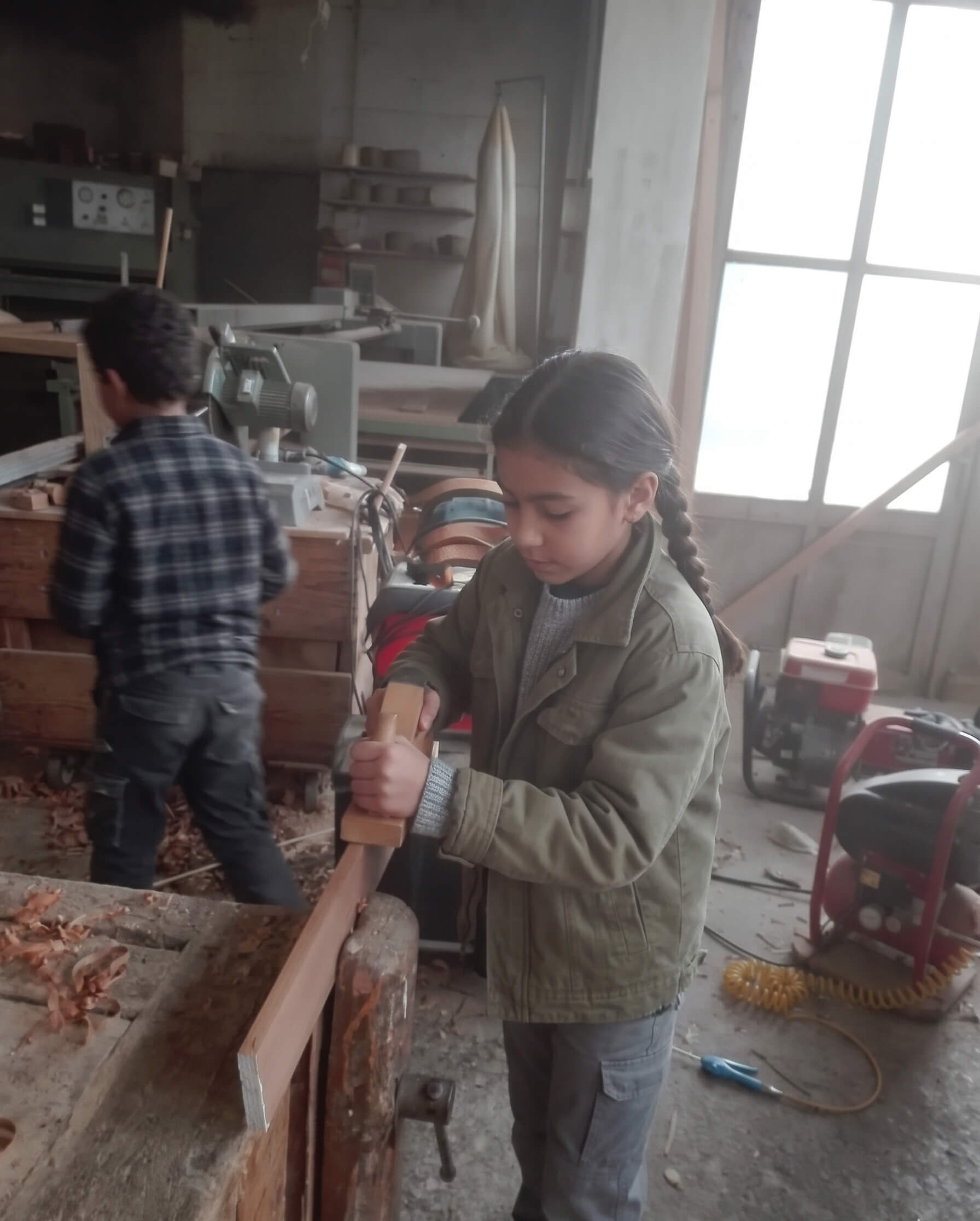 Bambina al banco da lavoro mentre rifinisce un pezzo di legno con un attrezzo manuale nel laboratorio.