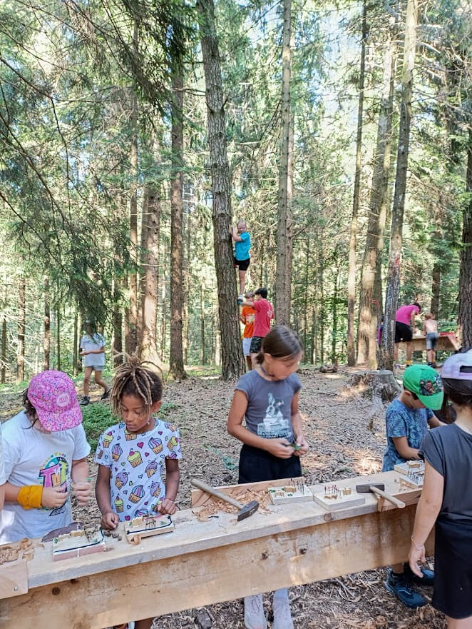 Bambini al lavoro su piccoli manufatti in legno in un laboratorio nel bosco con attivita' sugli alberi sullo sfondo.