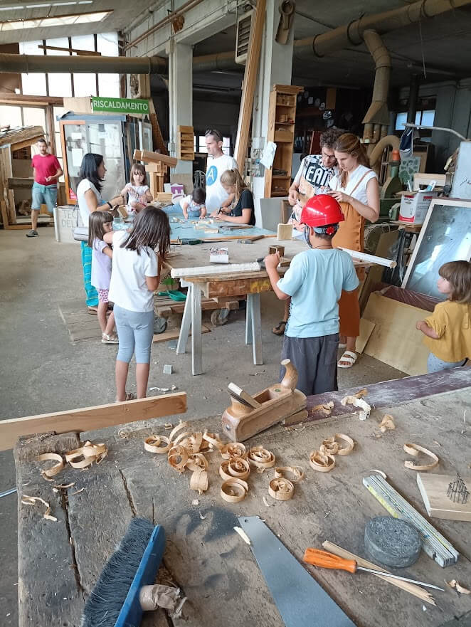 Laboratorio di falegnameria in ambiente interno con bambini al lavoro tra tavoli, attrezzi e trucioli di legno.