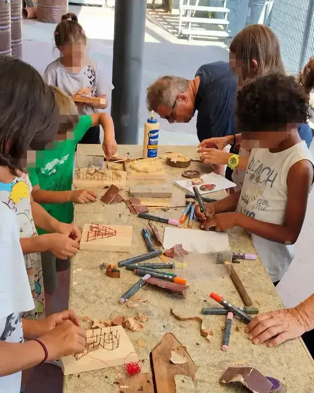 Bambini e adulti lavorano insieme attorno a un banco pieno di legni e strumenti.