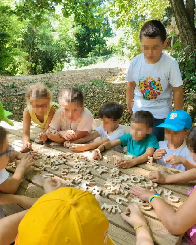 Bambini e un’adulta osservano lettere e forme disposte su un tavolo all’aperto.