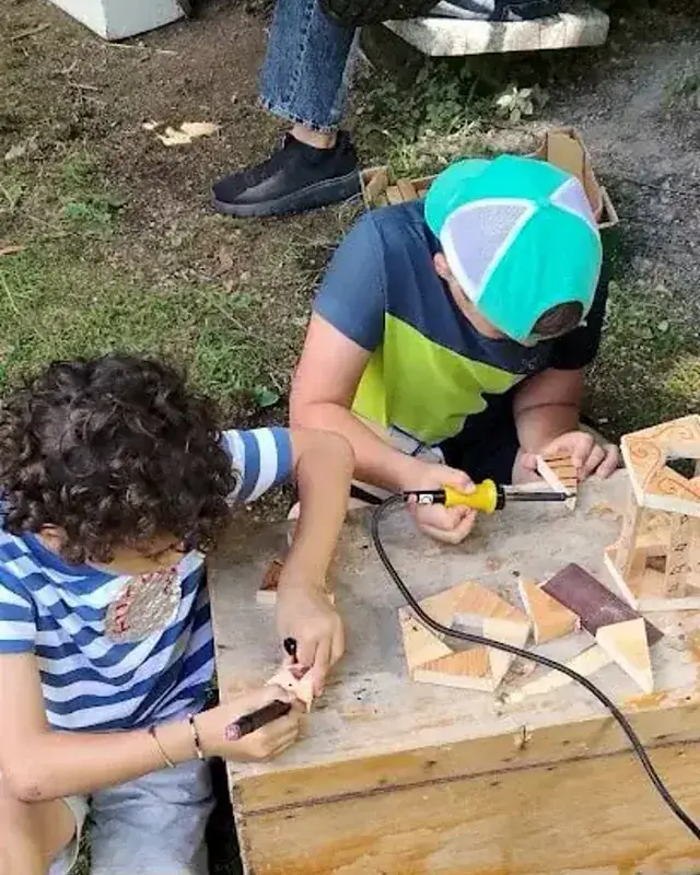 Due partecipanti lavorano piccoli pezzi di legno sul banco all’aperto.