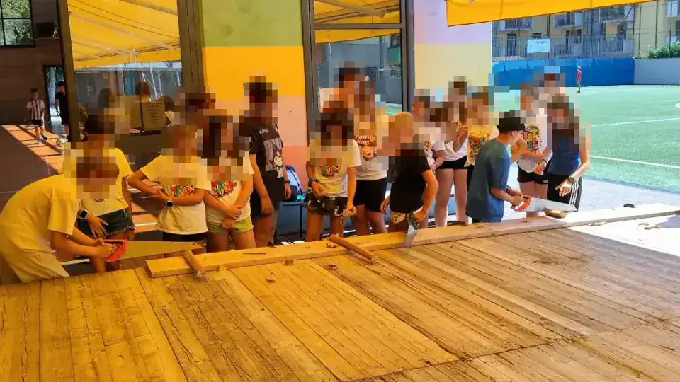 Un grande gruppo di ragazzi è disposto ai due lati del lungo banco di falegnameria.
