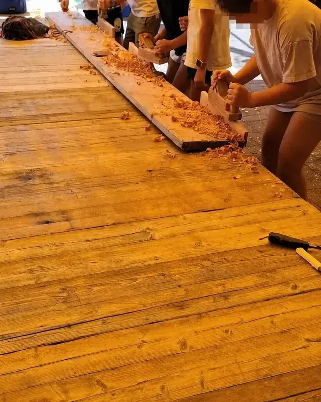 Una fila di ragazzi lavora il legno tra strumenti e trucioli sul banco.