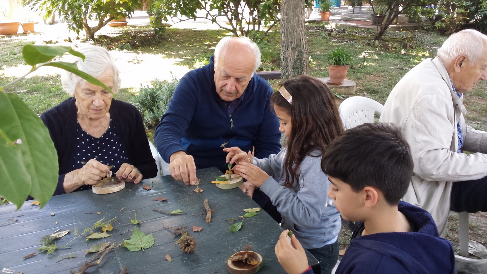 Persone anziane e bambini realizzano insieme piccoli lavori con materiali naturali in giardino.